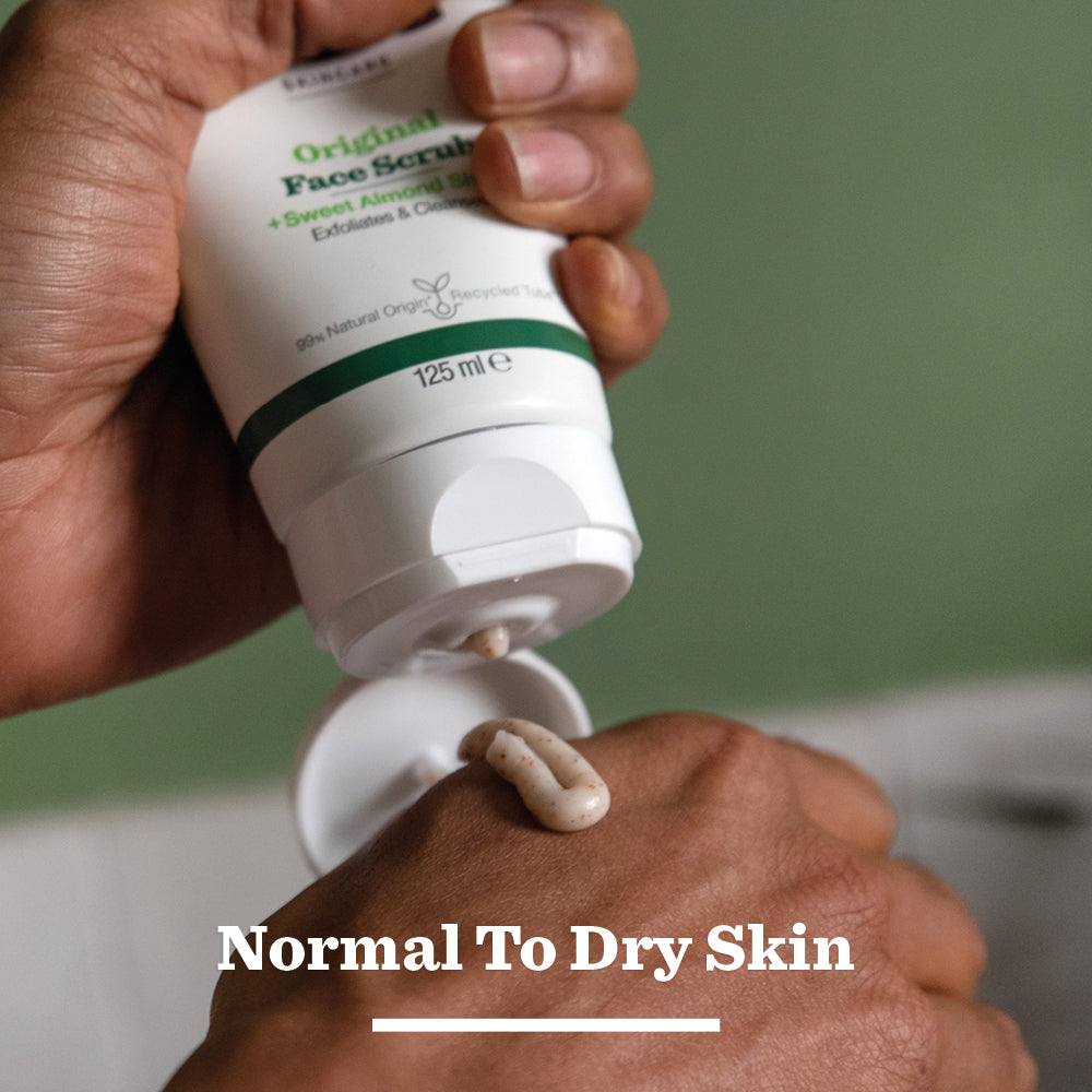 A man squeezes Original Face Scrub from a white tube onto his hand. Text highlights its exfoliating action with natural origin ingredients and suitability for normal to dry skin.