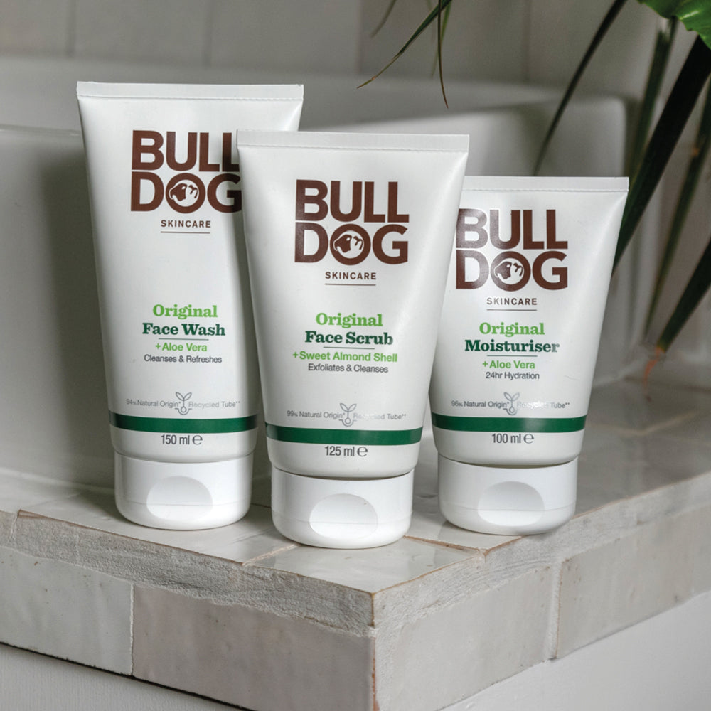 Three Bulldog Skincare products—Original Face Wash, Original Face Scrub, and Original Moisturiser—are displayed on a white bathroom counter with a plant in the background.