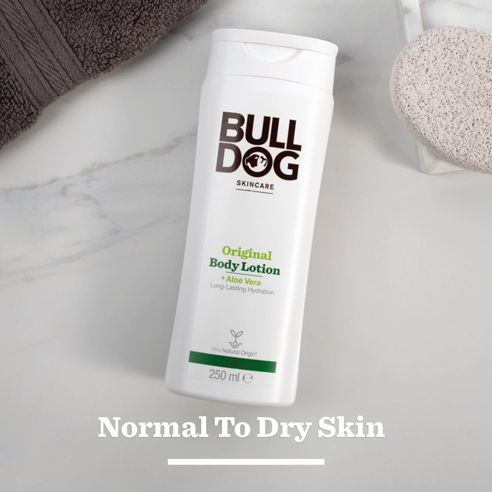 A white bottle of Original Body Lotion is placed on a marble surface beside a brown towel and a round exfoliating sponge. Normal To Dry Skin appears at the bottom of the image.