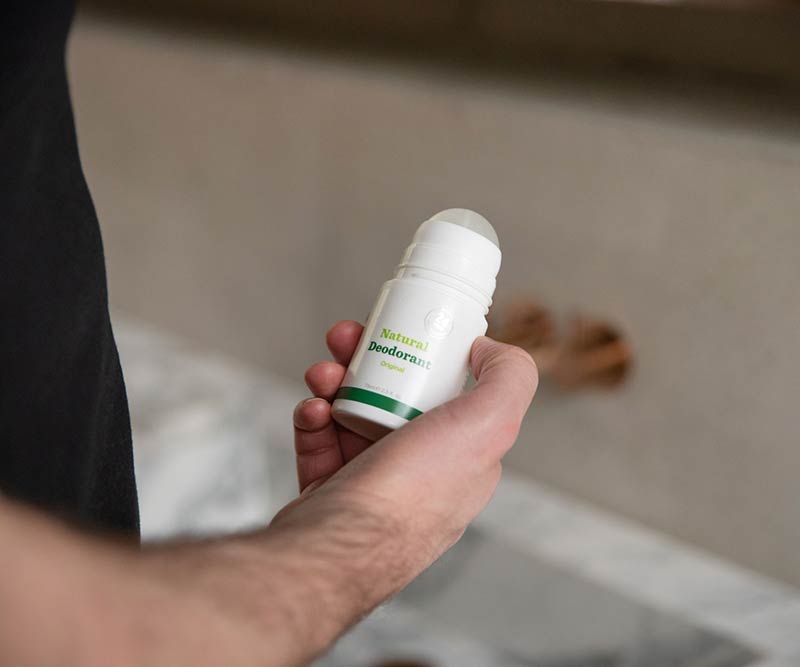 A person holds a white container from the Premium Body Care Bundle, labeled Natural Deodorant with a green stripe and aloe vera, standing near a bathroom sink.