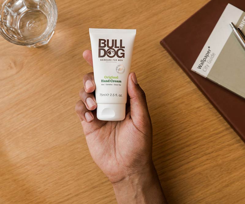 A hand holds a tube of Bulldog Original Hand Cream with aloe vera over a wooden table, next to a water glass and brown notebook—ideal companions for your Premium Body Care Bundle essentials.